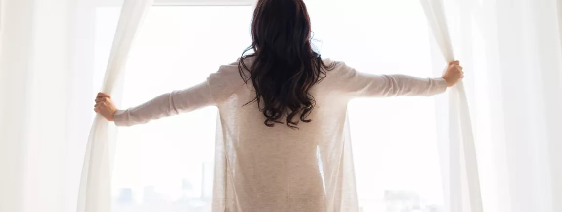 Comment garder son intérieur frais tout l’été ? Une femme ouvre les rideau d’une fenêtre d’appartement